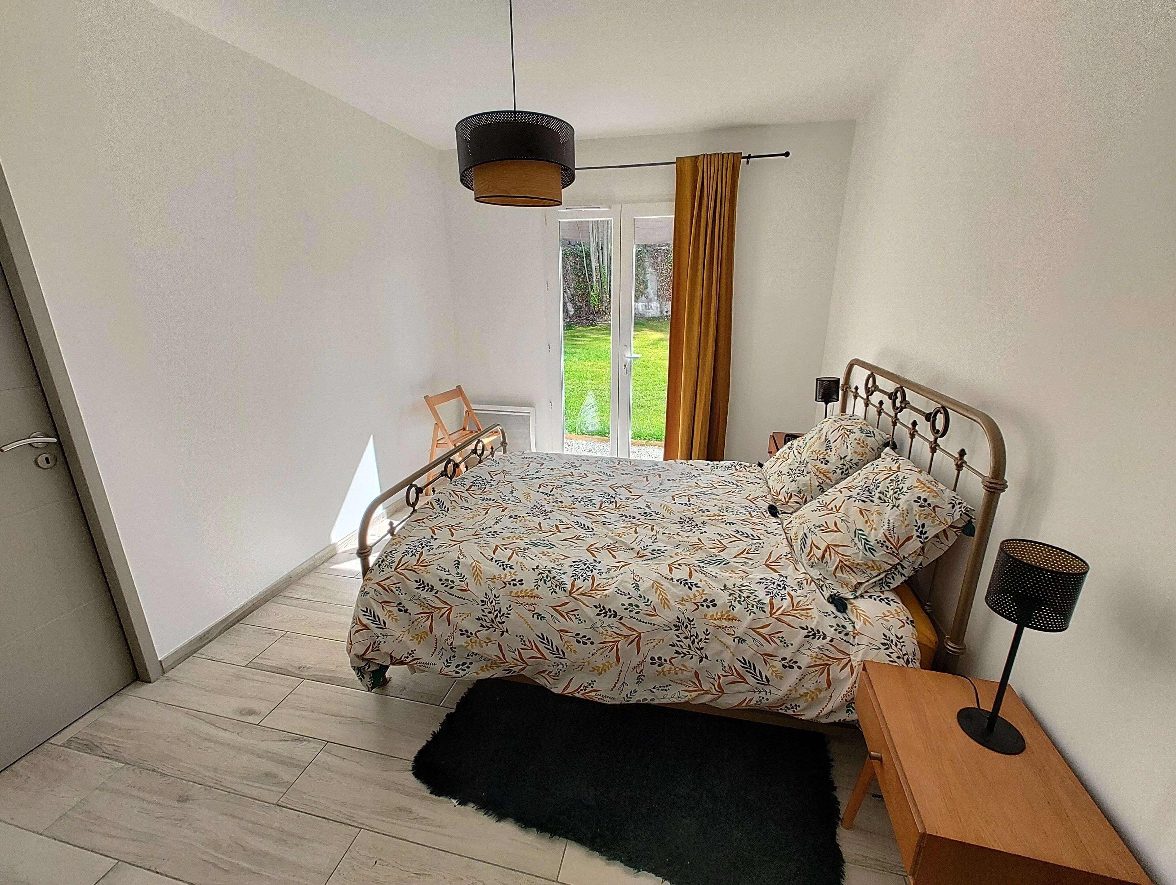A bedroom with a bed, a black rug, a wooden nightstand, a lamp, and a view of a garden through a sliding door.