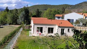 A modern two-story house with a red-tiled roof, a small patio with outdoor furniture, and a view of a lush green landscape and mountains in the background. - Maison de Vacances Plain Pied 6 Couchages -2 sdb - Jardin -vernet les Bains (Vernet-les-Bains)