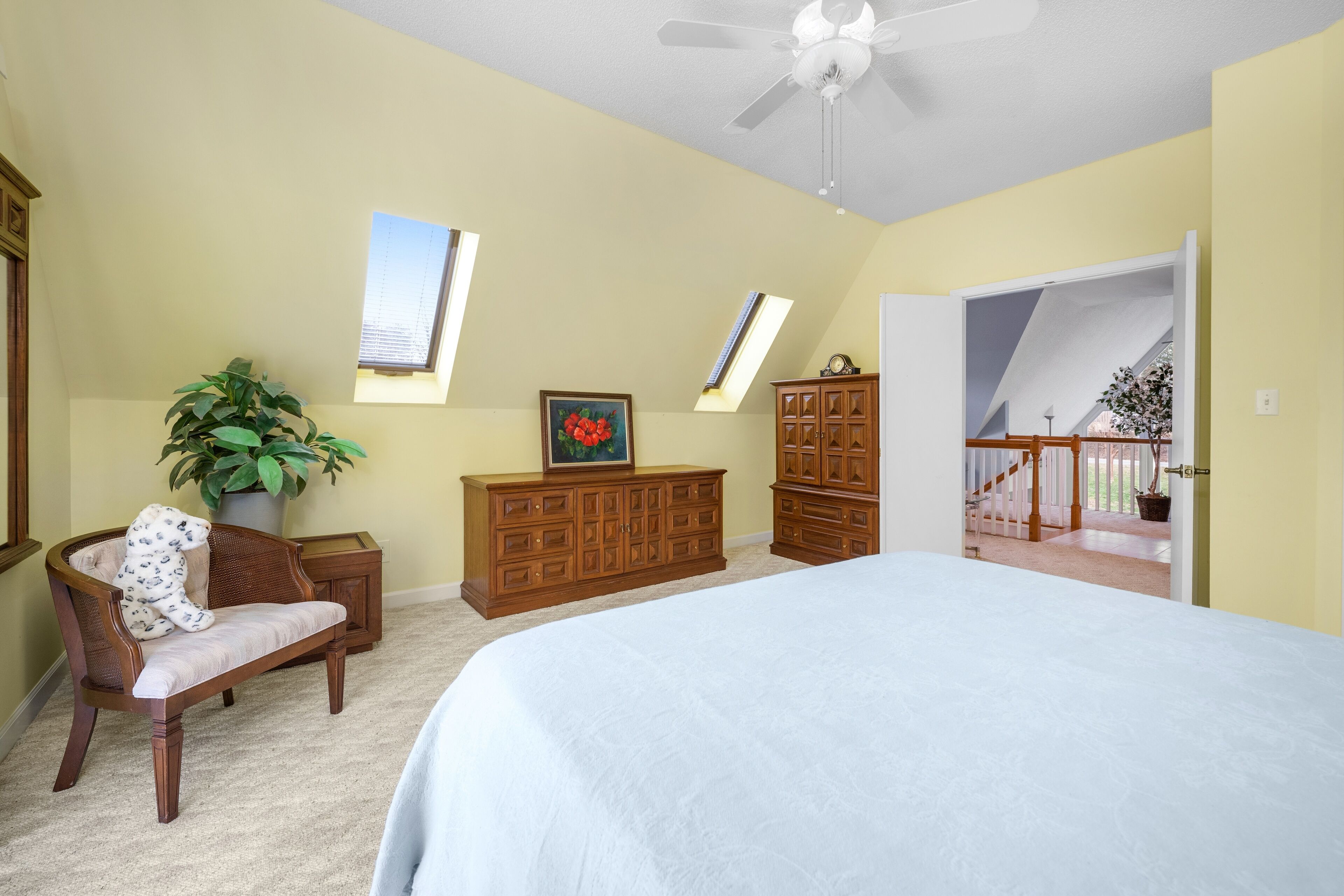 A bedroom with a large bed, a wooden dresser, a chair, a potted plant, and a ceiling fan.