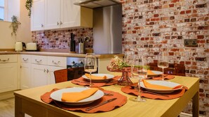 Un comedor con pared de ladrillo, armarios blancos y una mesa dispuesta para dos personas, con manteles individuales naranja y copas de vino.
