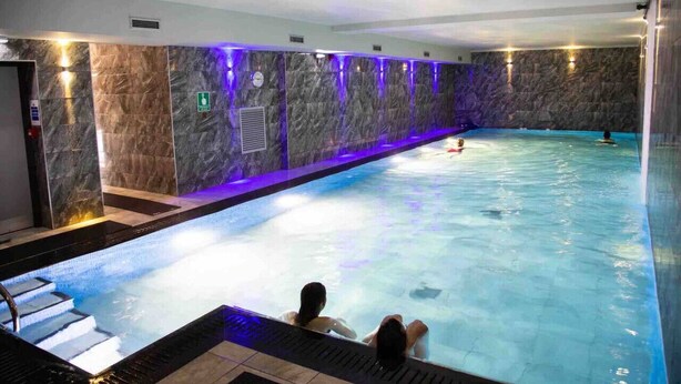 Una piscina interior con pared de mármol, una persona relajándose en el borde y otra nadando.