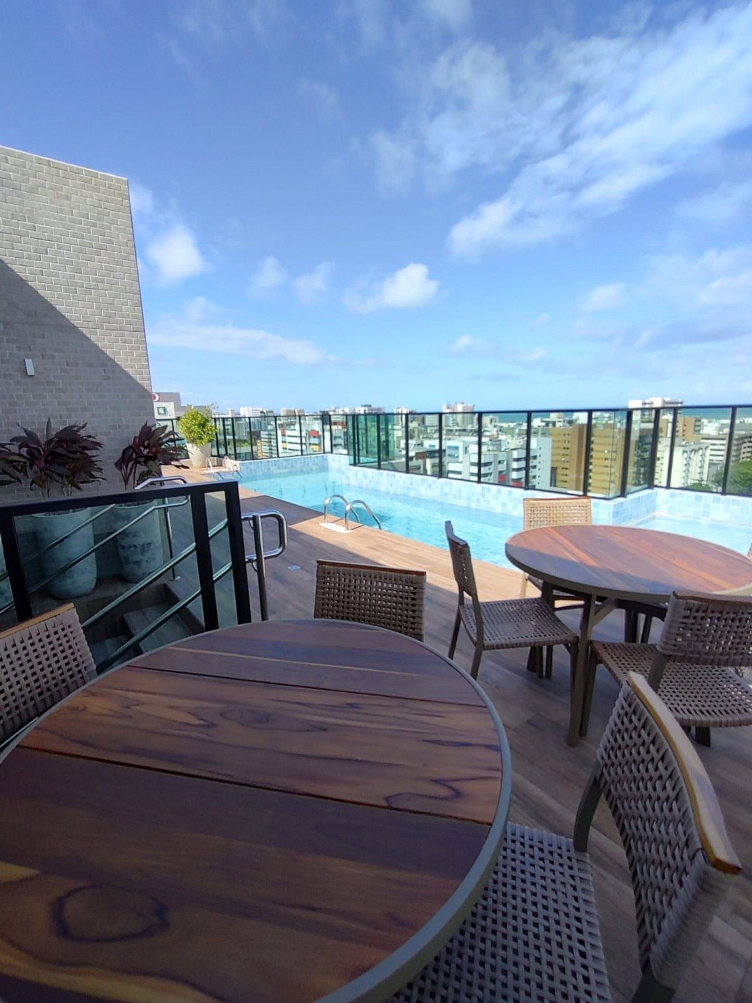 A rooftop pool area with a wooden table and wicker chairs, surrounded by glass railings and potted plants.