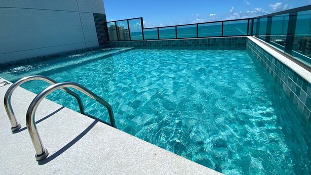 A clear blue pool with a metal ladder on a tiled deck, surrounded by a glass railing and a white wall. - Edificio Liv P.Verde Apto 1108 (Maceió)
