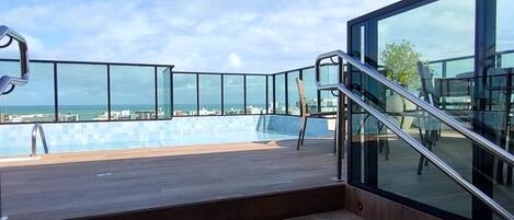 A modern outdoor pool area with a view of the sea, wooden steps, and glass railings.