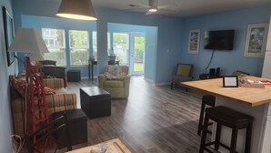 A spacious living area with a wooden floor, a sofa, a chair, a coffee table, and a dining table with stools. - Cozy Pool side Condo, 5 min from the beach! (Little River)