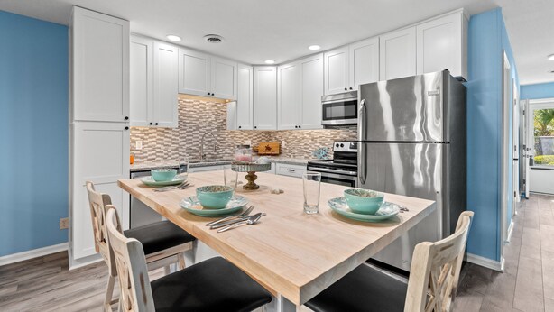 A modern kitchen with white cabinets, a stainless steel refrigerator, and a wooden dining table with teal bowls. - Cozy Pool side Condo, 5 min from the beach! (Little River)