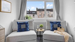 A cozy room with a grey sofa set, a small table with a plant, and a view of buildings through a window with grey curtains. - Gloster Town House Gorgeous Family Home Barnstaple (Barnstaple)