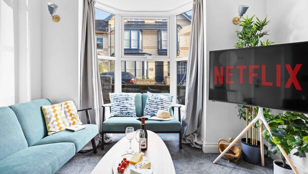A modern living room with a large window, a sofa, a coffee table with snacks and drinks, and a TV screen displaying "NETFLIX". - Gloster Town House Gorgeous Family Home Barnstaple (Barnstaple)