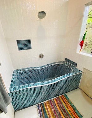 A bathtub with mosaic tiles, a window with a plant, and a colorful striped rug.
