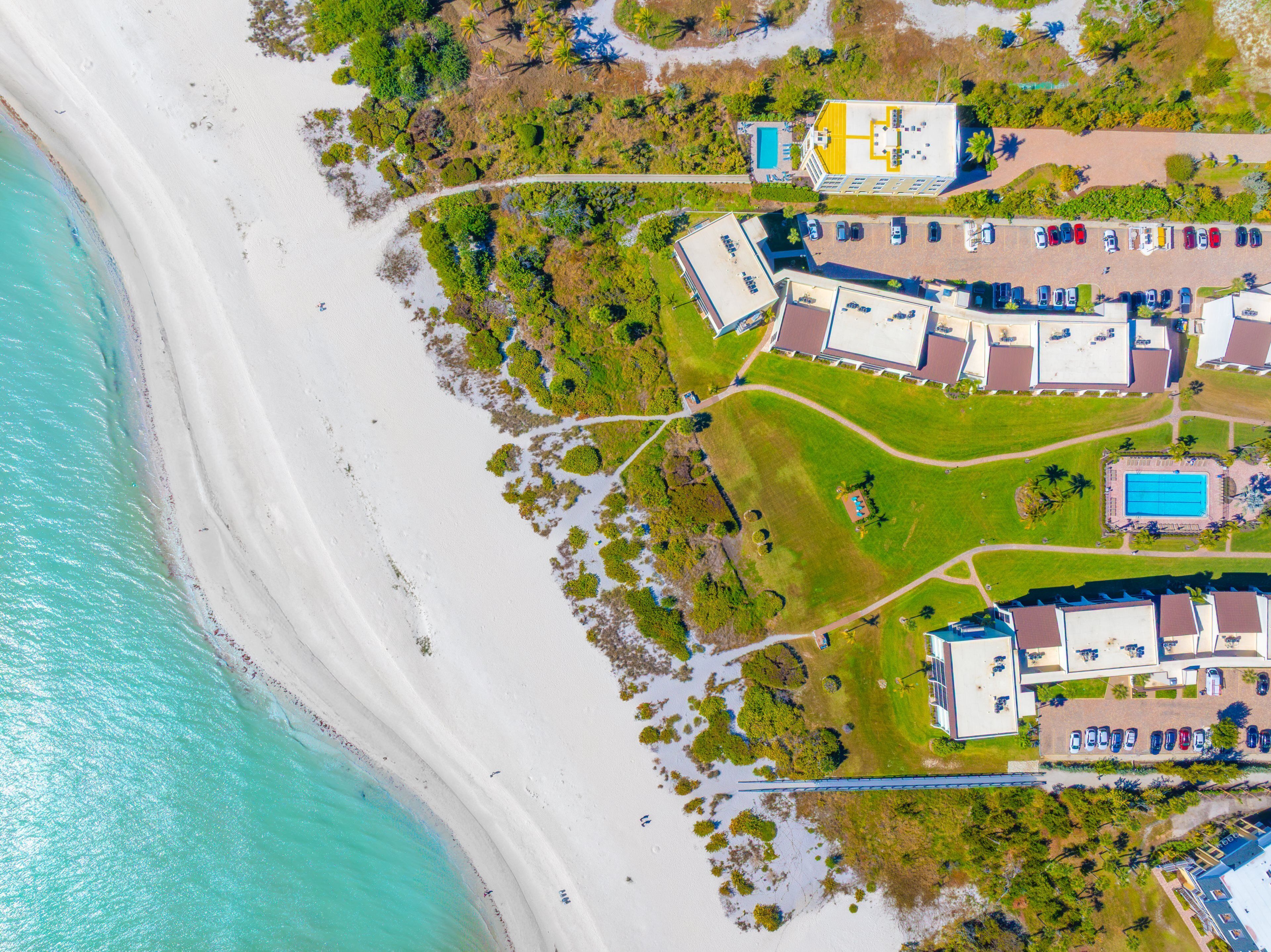 An aerial view of a coastal resort with a beach, swimming pools, and multiple buildings with parking areas.