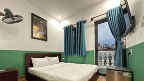 A hotel room with a bed, a desk, a chair, a window with curtains, a radiator, and a wall-mounted fan. - Nineties Homestay  (Hue)