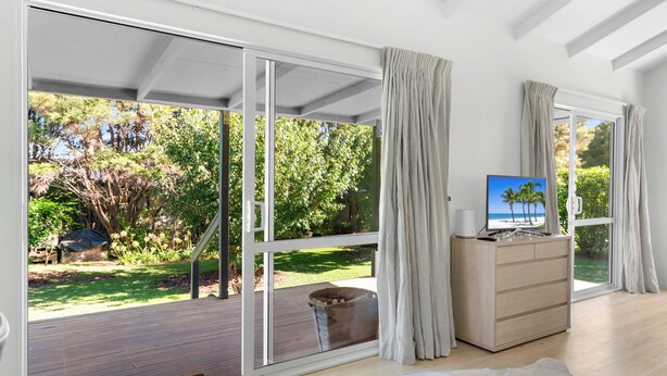 Ein modernes Zimmer mit einer großen Schiebeglastür, die auf eine Terrasse führt, einem Holzkoffer und einem Fernseher, der eine Strandansicht zeigt.