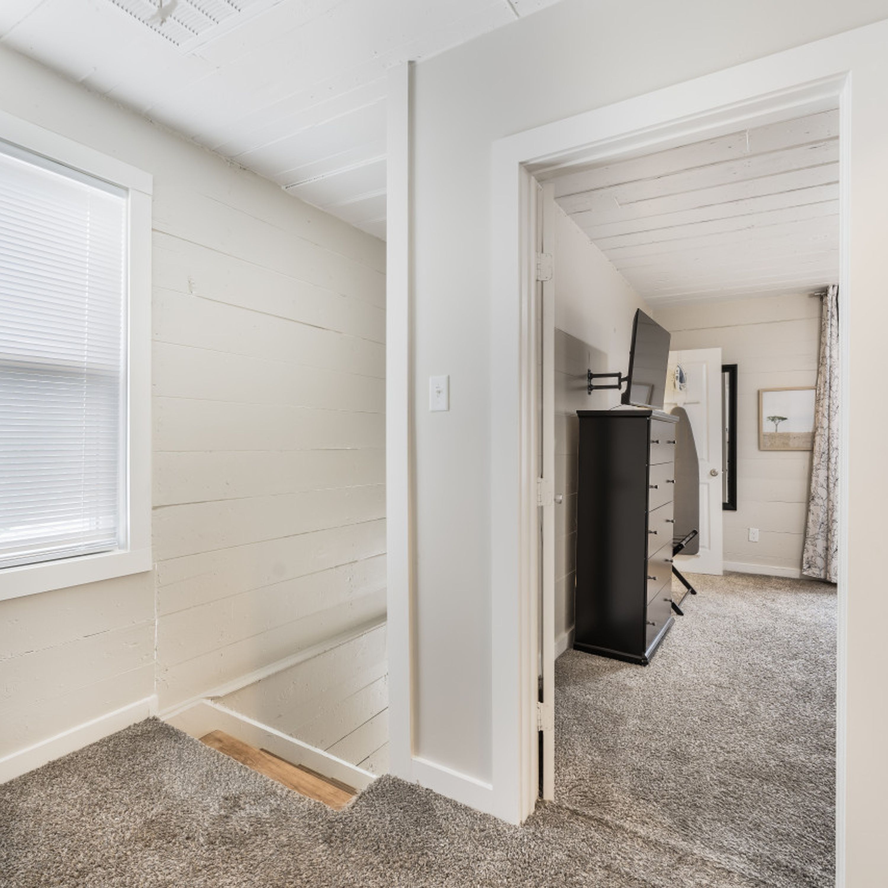 A room with a sloped ceiling, a window with blinds, a door leading to another room, and a carpeted floor.