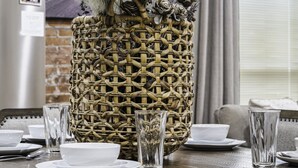 Une table à manger est dressée avec de l’assiette blanche, des bols et de la verrerie transparente. Un panier tressé contenant des fleurs séchées et des branches est posé sur la table.