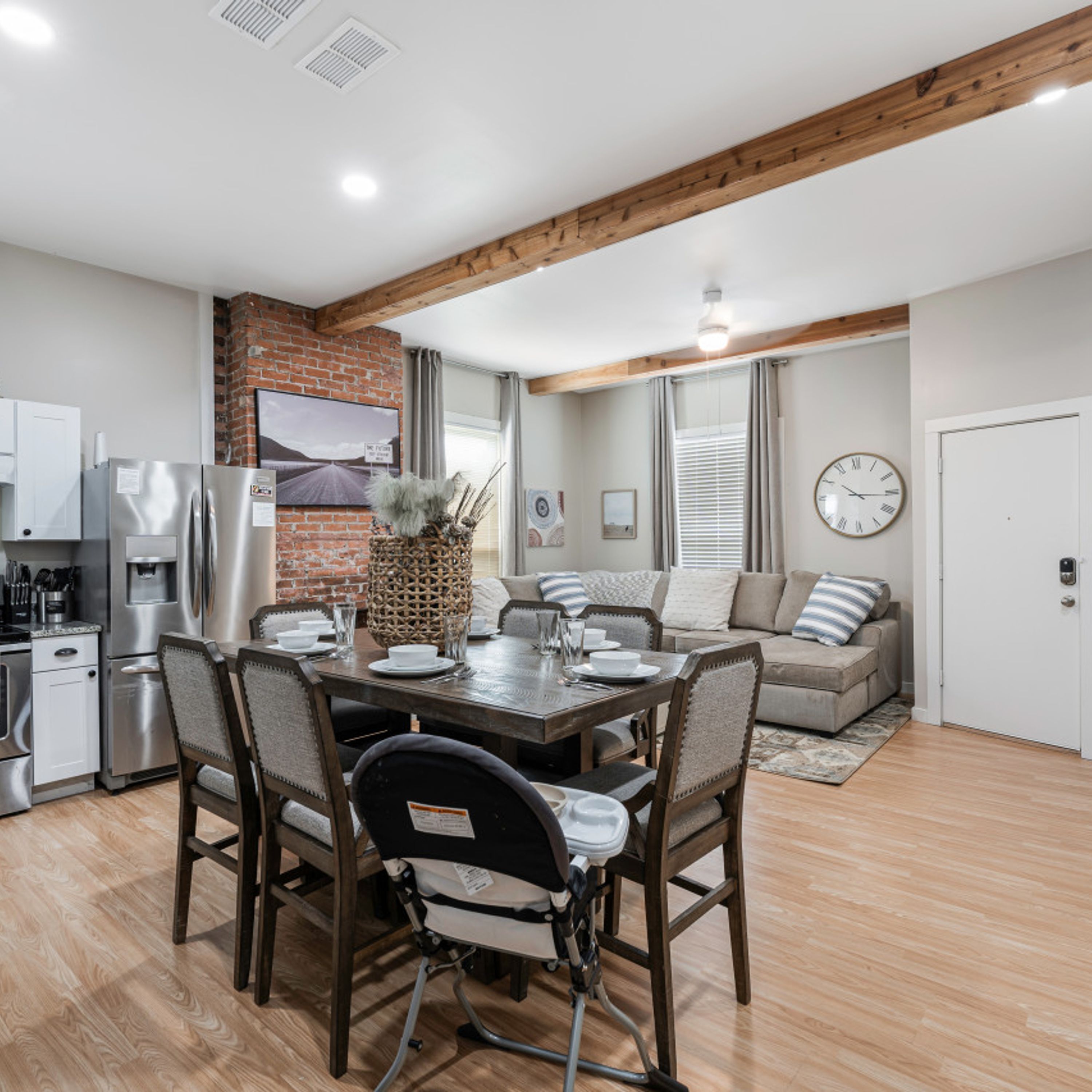 A modern kitchen with stainless steel appliances, a dining area with a wooden table and chairs, and a living area with a sofa and a clock on the wall.