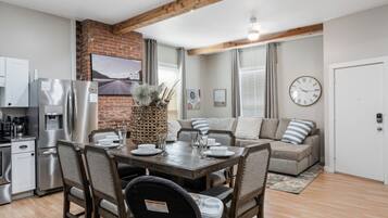 A modern kitchen with stainless steel appliances, a dining area with a wooden table and chairs, and a living area with a sofa and a clock on the wall.