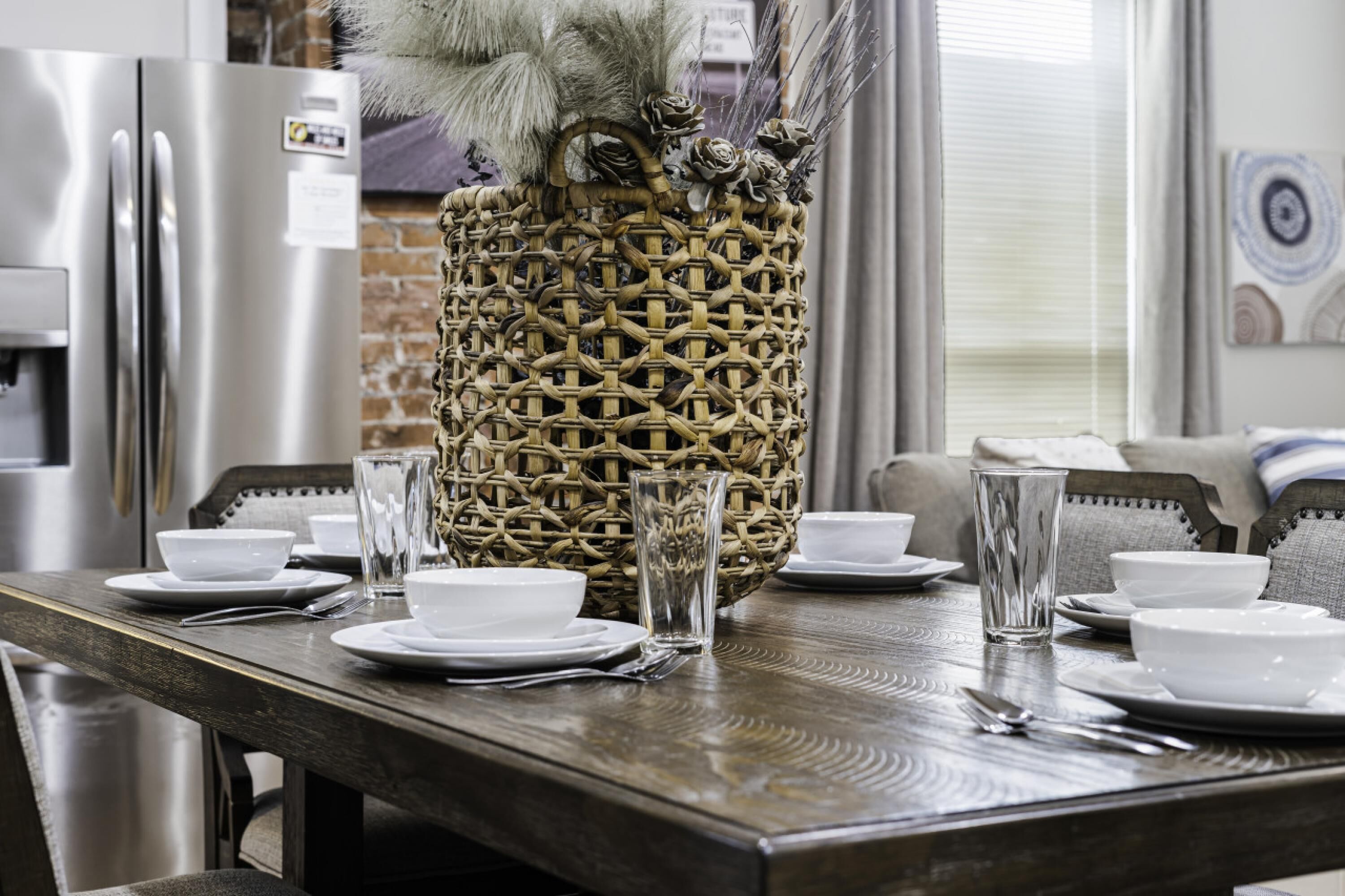 A dining table set with white plates, bowls, and clear glasses, with a woven basket containing dried flowers in the center.
