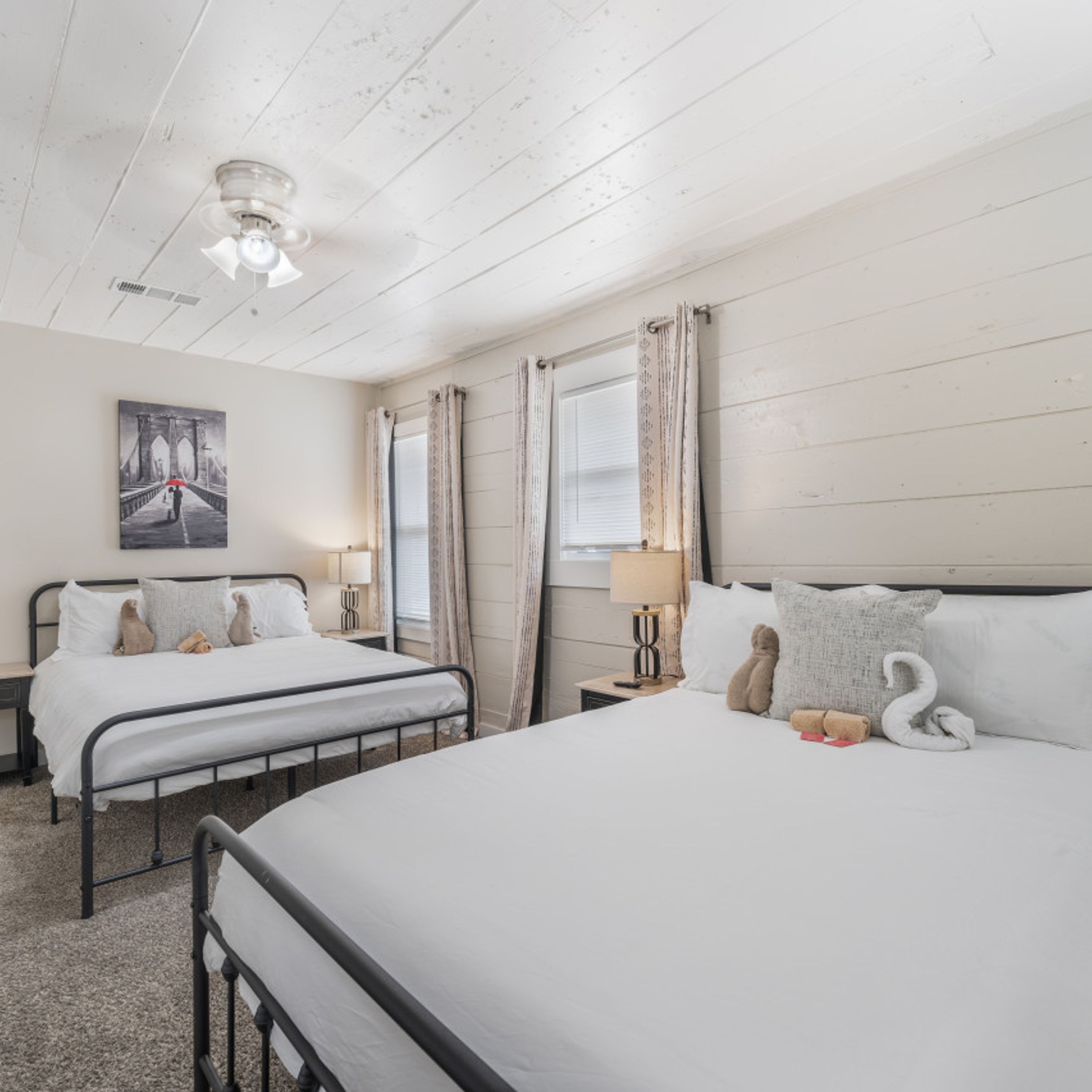 A hotel room with two beds, a ceiling fan, a painting of a bridge, and a window with curtains.