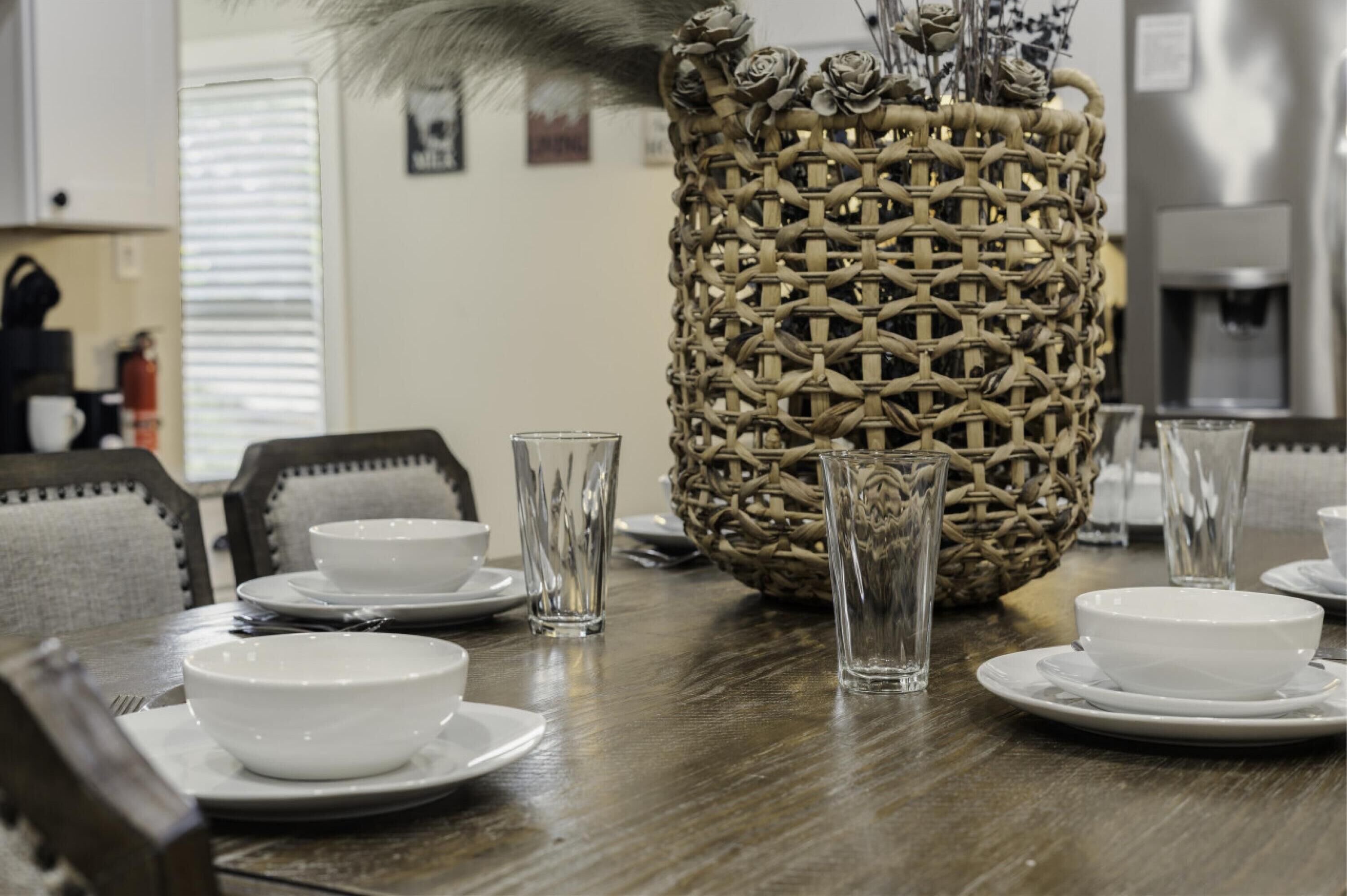 A dining table set with white bowls and clear glasses, a woven basket, and a decorative vase with dried flowers.
