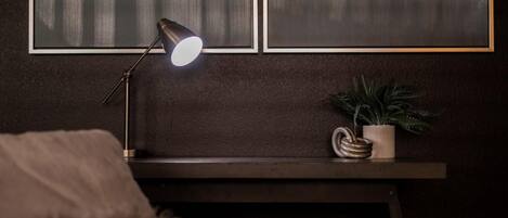 Ein dunkles Zimmer mit Schreibtisch, Lampe, Pflanze und gepolstertem Sitzplatz.