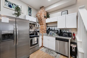 A modern kitchen with stainless steel appliances, white cabinetry, a brick oven, and a backsplash with hanging pots. - 4 Queen Beds High Speed Wifi (Fort Worth)
