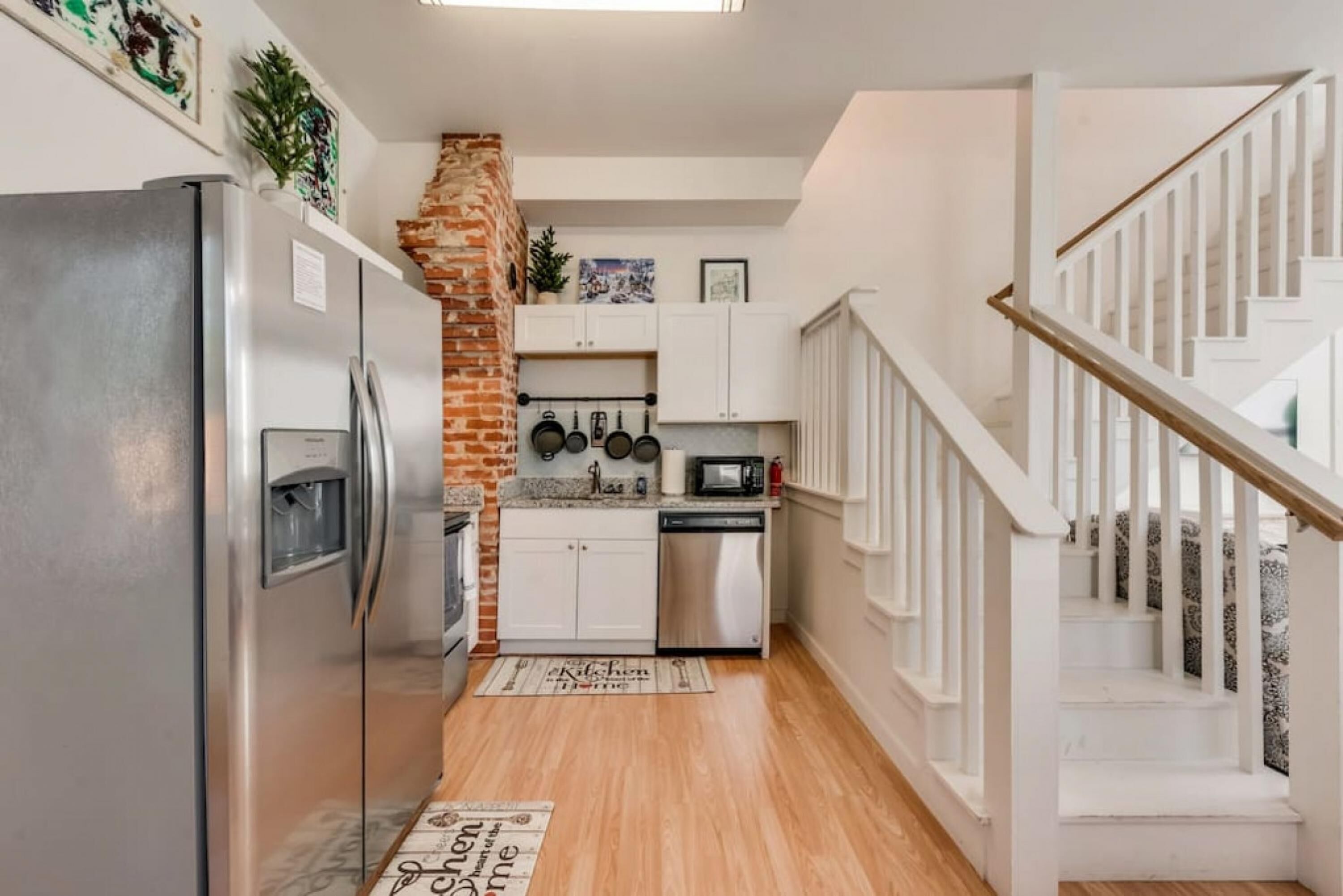 Cocina compacta con refrigerador, microondas y estufa, pared de ladrillo, gabinetes blancos y una escalera de madera.