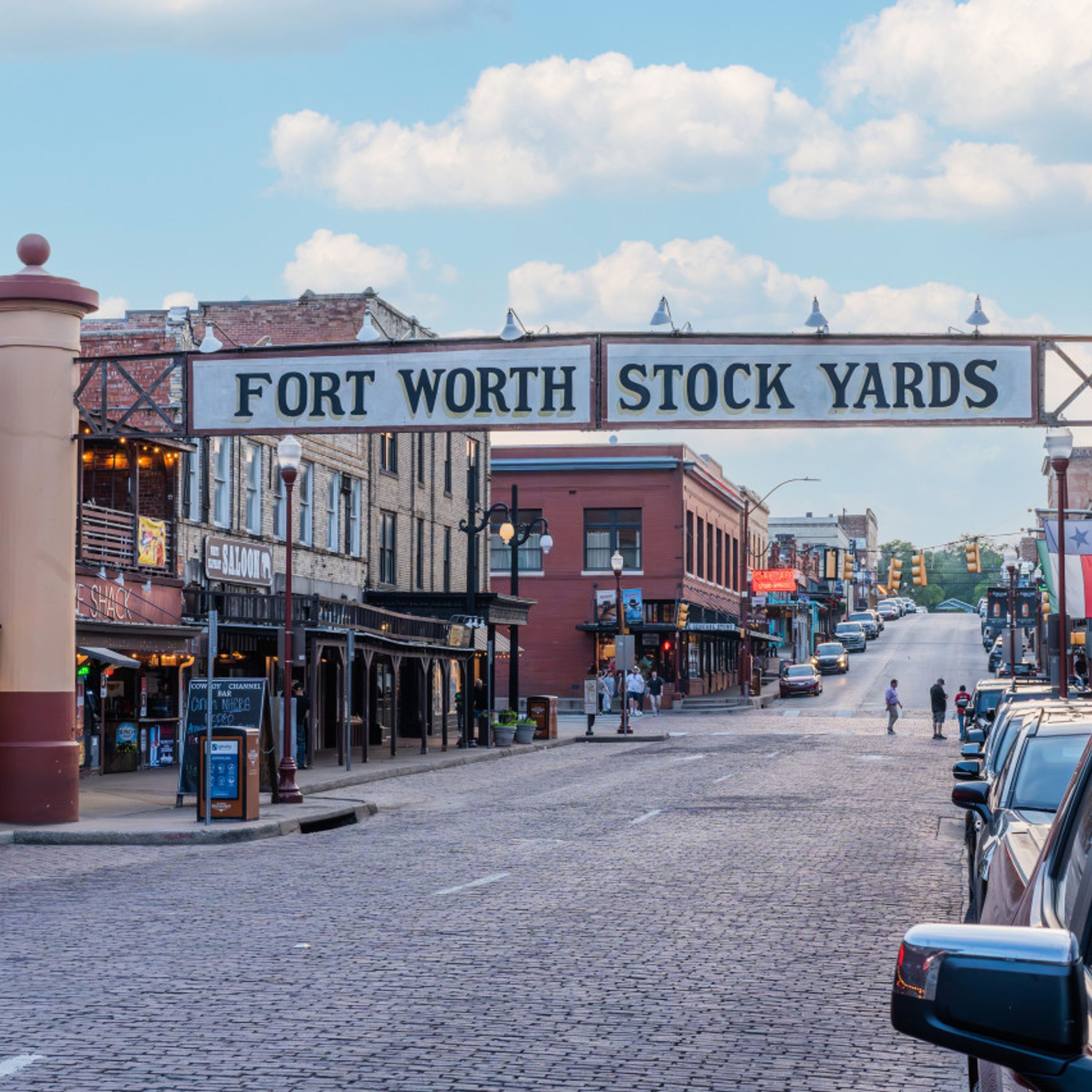 Una calle adoquinada con edificios a ambos lados, un letrero que dice "FORT WORTH STOCK YARDS" y coches estacionados.