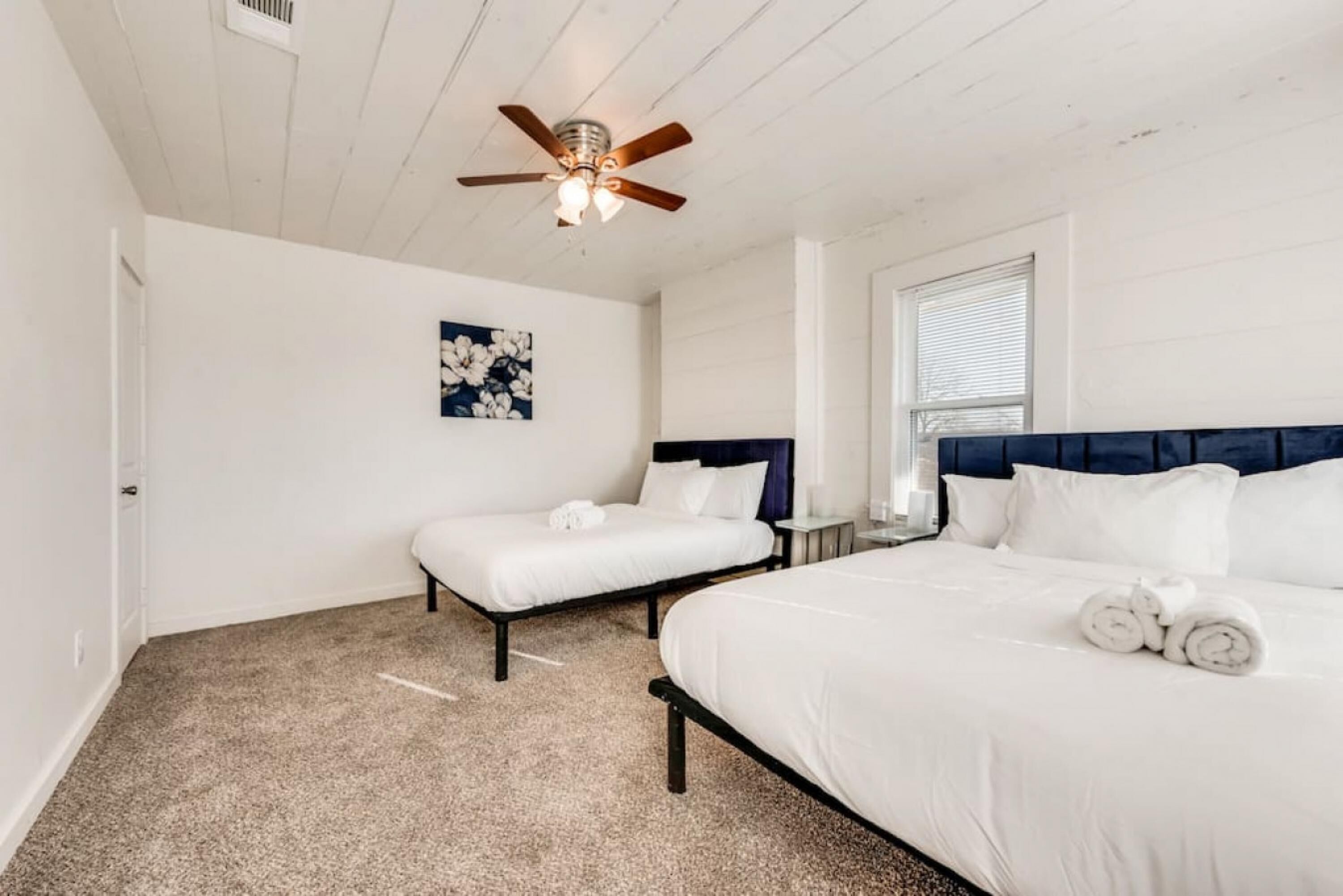 A bedroom with two beds, a ceiling fan, a window with blinds, and a painting on the wall.