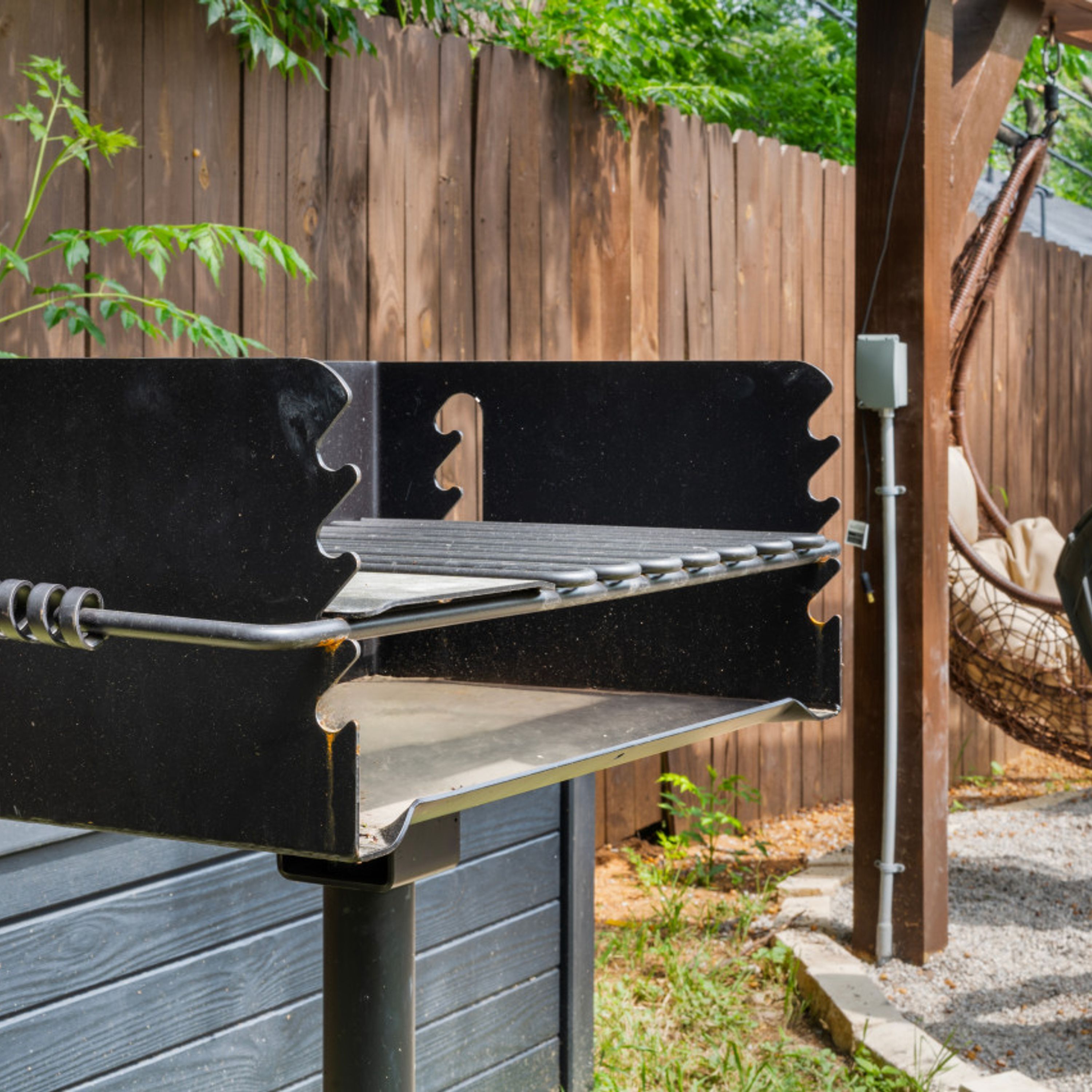 Un barbecue doté d’une structure métallique et d’une surface de cuisson plane, installé à l’extérieur, avec une clôture en bois et de la verdure en arrière-plan.
