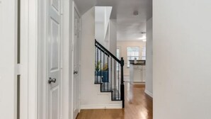 A hallway with wooden flooring, white walls, and a black staircase leading to a kitchen area. - Family Business Retreat King Bed Top Amenities (Euless)
