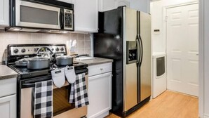 A kitchen with white cabinets, a black refrigerator, a microwave, a stove, and a checkered oven mitts hanging on the oven handle. - Family Business Retreat King Bed Top Amenities (Euless)