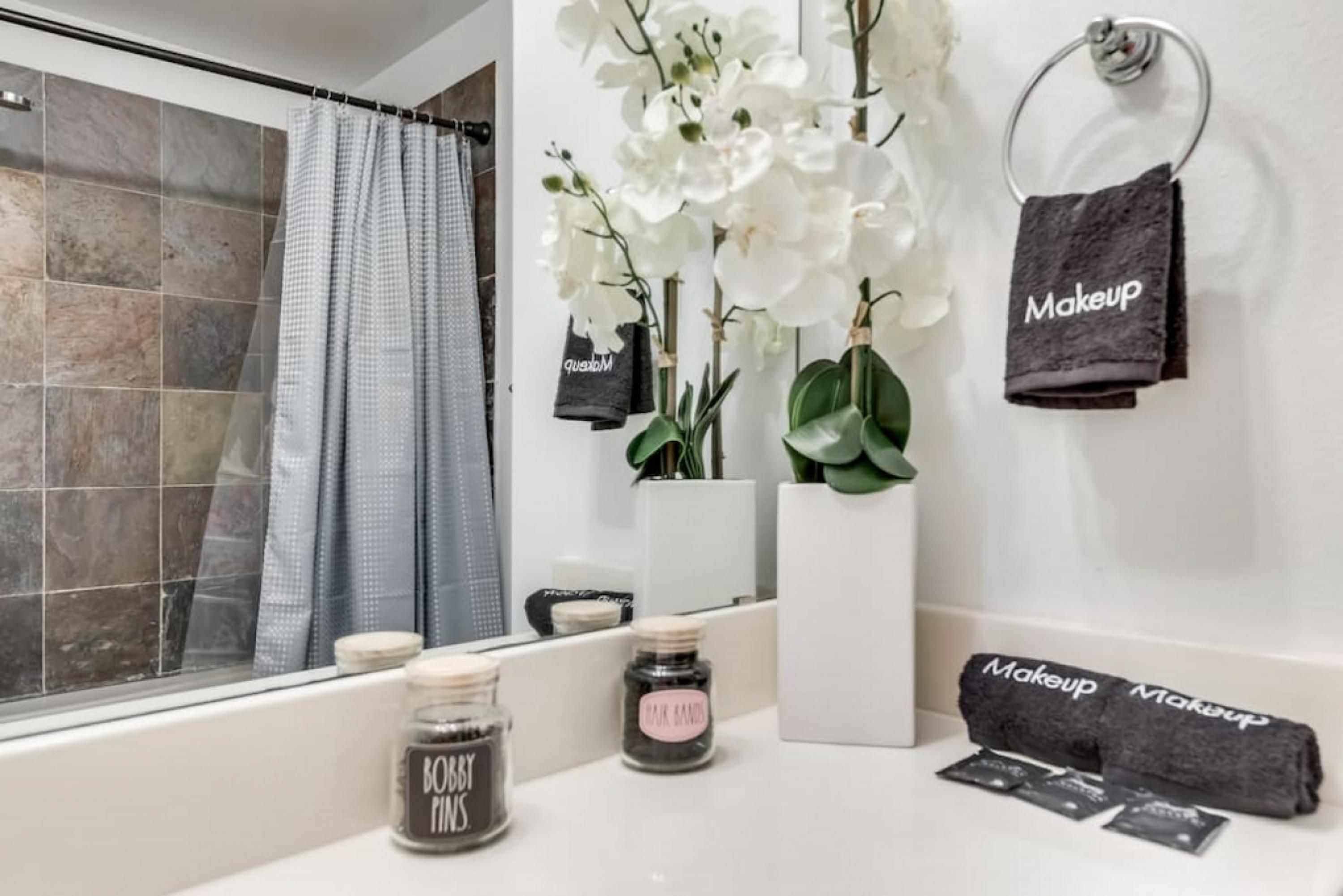 A bathroom with a bathtub, a shower curtain, a towel rack with a towel, a vase with white flowers, and two jars labeled "BOBBY RINGS" and "MAKEUP".