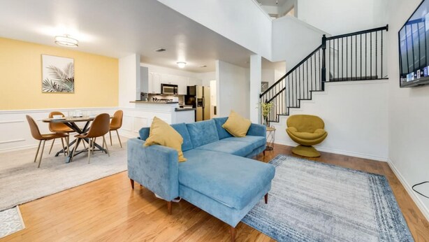 A modern living room with a blue sectional sofa, a dining table with chairs, a staircase, and a kitchen in the background. - Family Business Retreat King Bed Top Amenities (Euless)