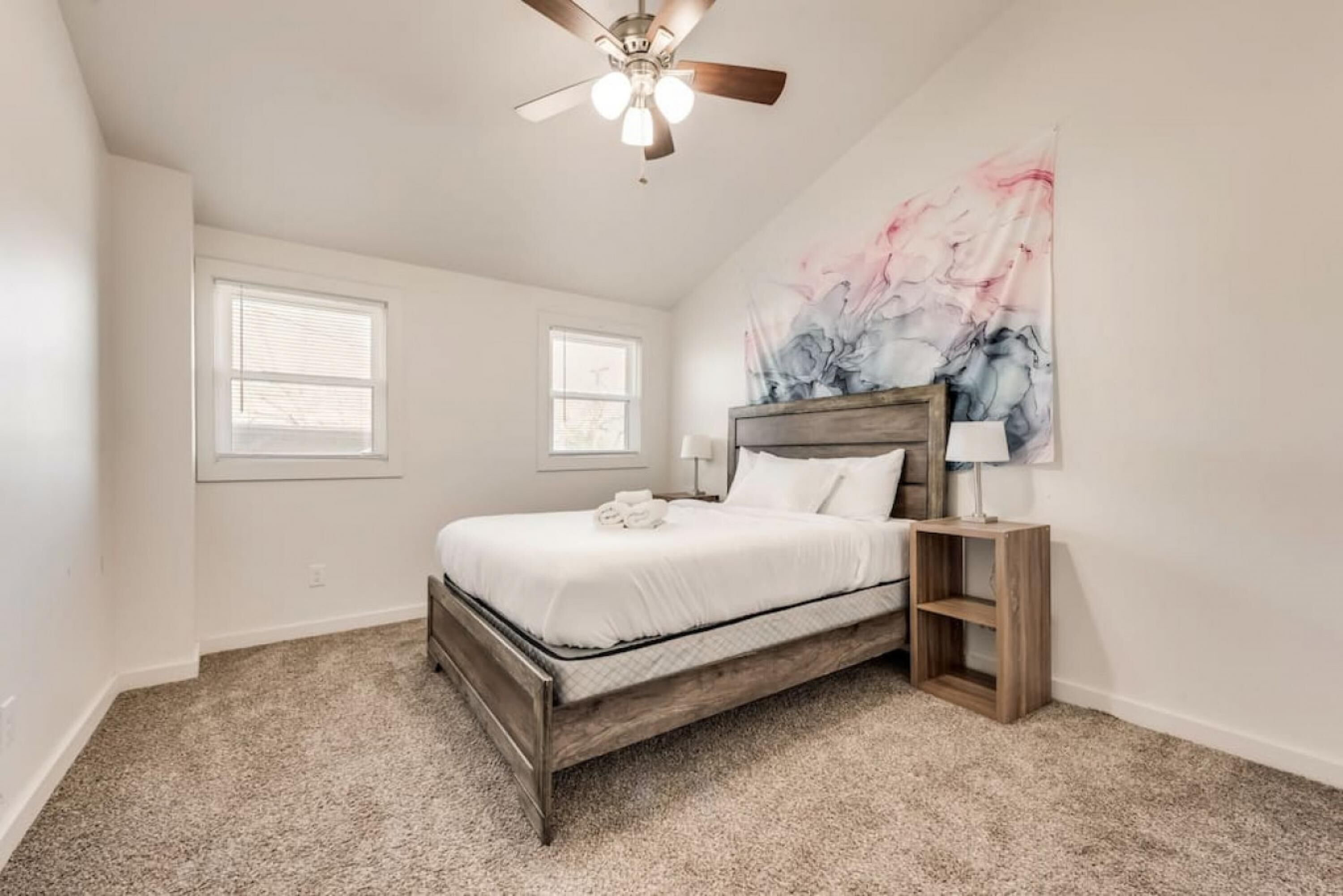Ein Schlafzimmer mit einem Bett, Deckenventilator, zwei Fenstern und einem Wandbild.