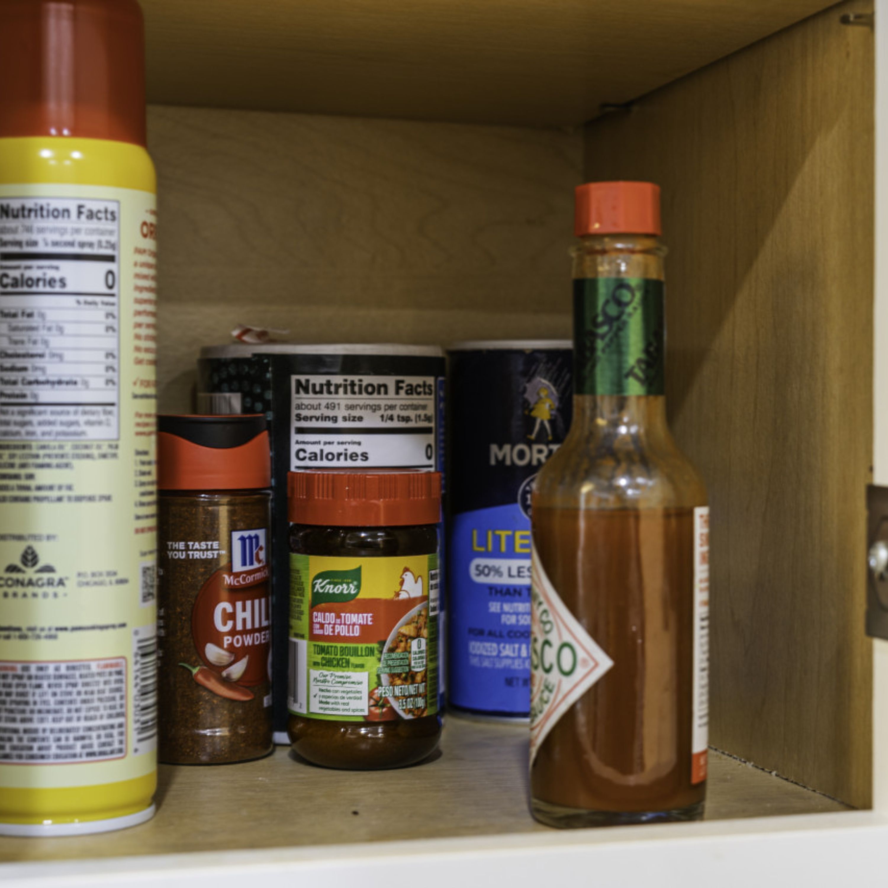 Una repisa con varios condimentos, incluyendo una botella de salsa picante, un frasco de chile en polvo y un envase de salsa de tomate Knorr.