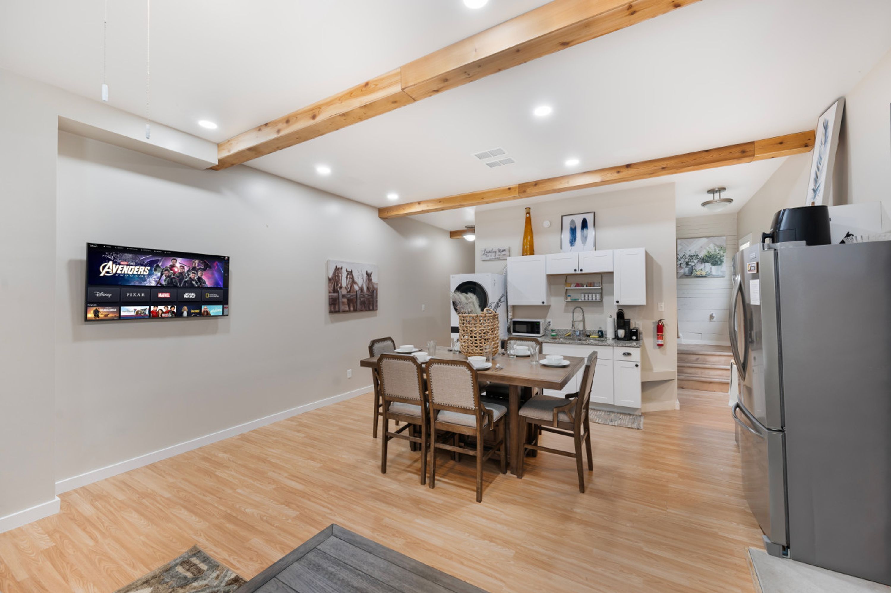 A modern kitchen with wooden beams, a dining area with a table and chairs, and a flat-screen TV displaying the Avengers movie.