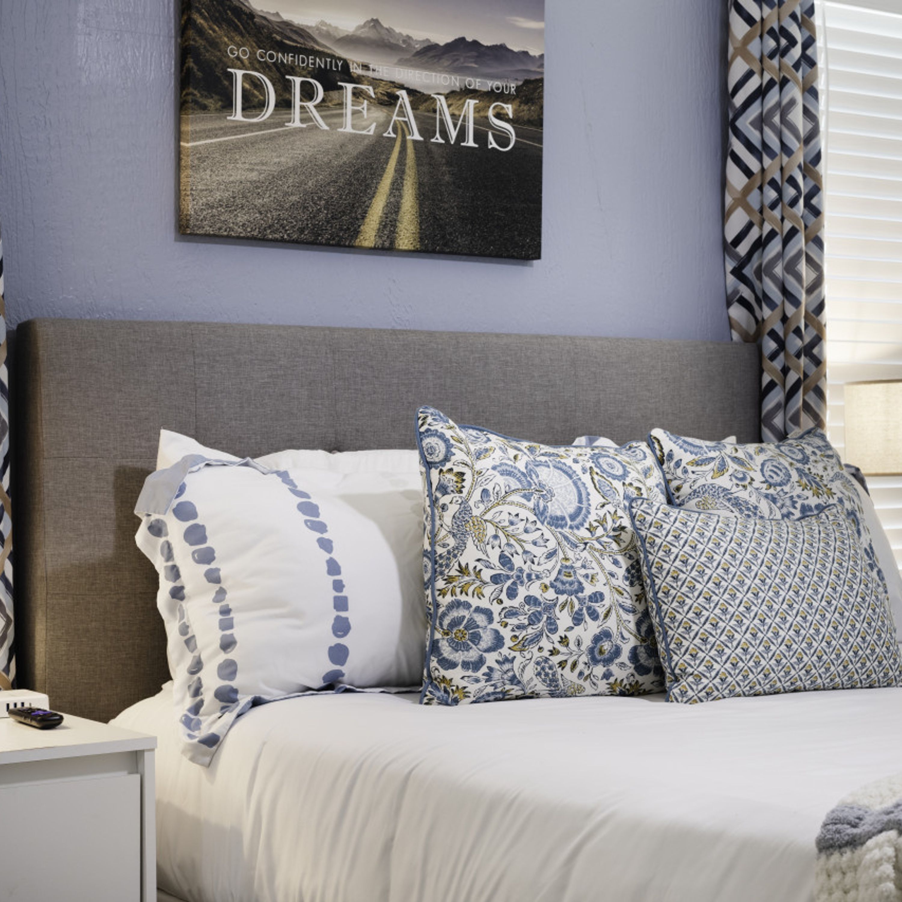 A neatly made bed with patterned pillows, a framed picture on the wall, and a patterned curtain.