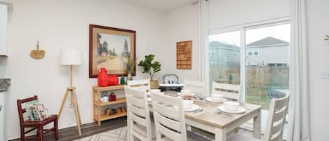 A dining area with a table set for four, surrounded by white chairs, a red object on a shelf, and a painting on the wall.
