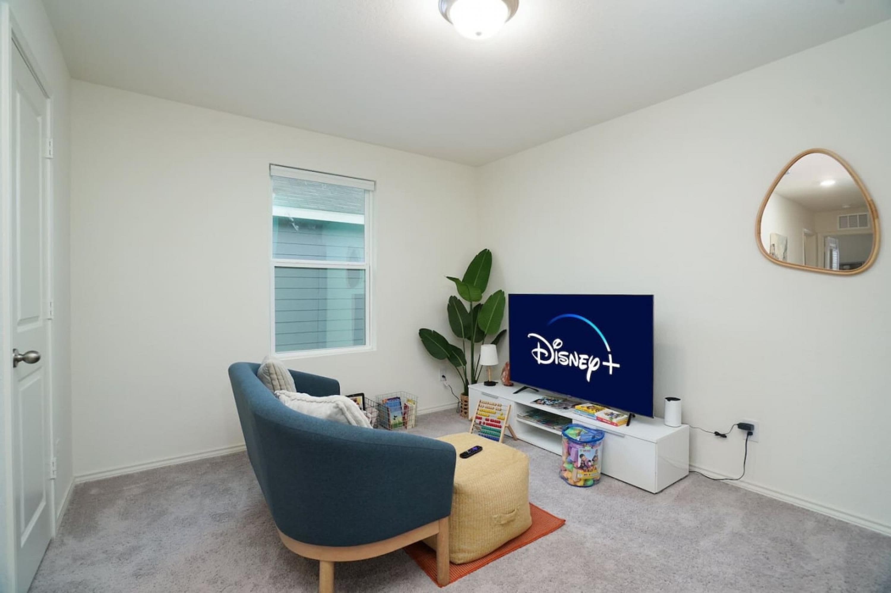 Un salon chaleureux avec un fauteuil bleu, un téléviseur à écran plat diffusant Disney+, une petite table avec des livres et une plante, et un miroir au mur.