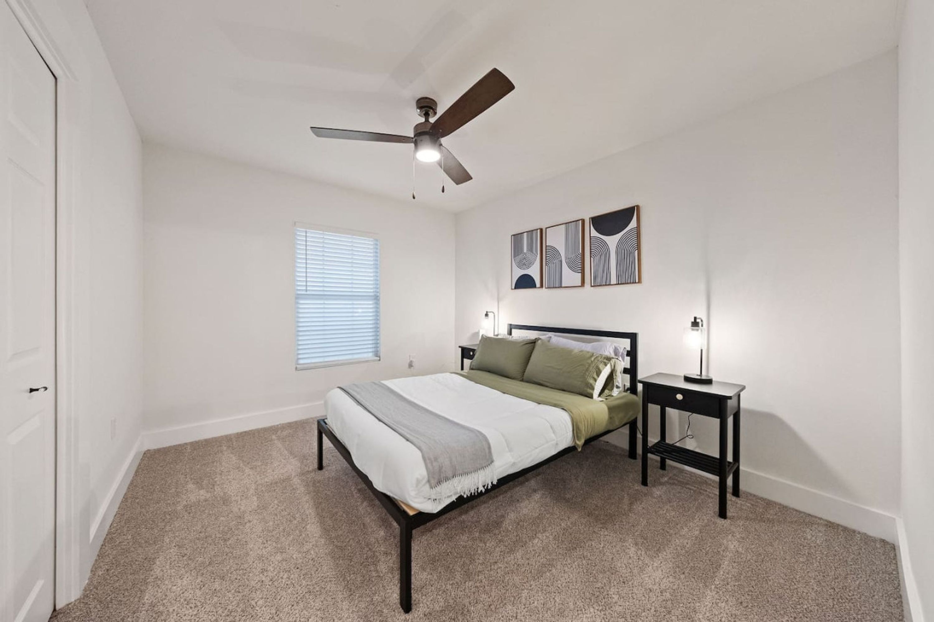 A bedroom with a bed, a nightstand, a ceiling fan, a window with blinds, and wall art.