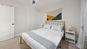 Un dormitorio con una cama, un ventilador de techo, una puerta blanca, una mesita de noche blanca y un tapiz en la pared.