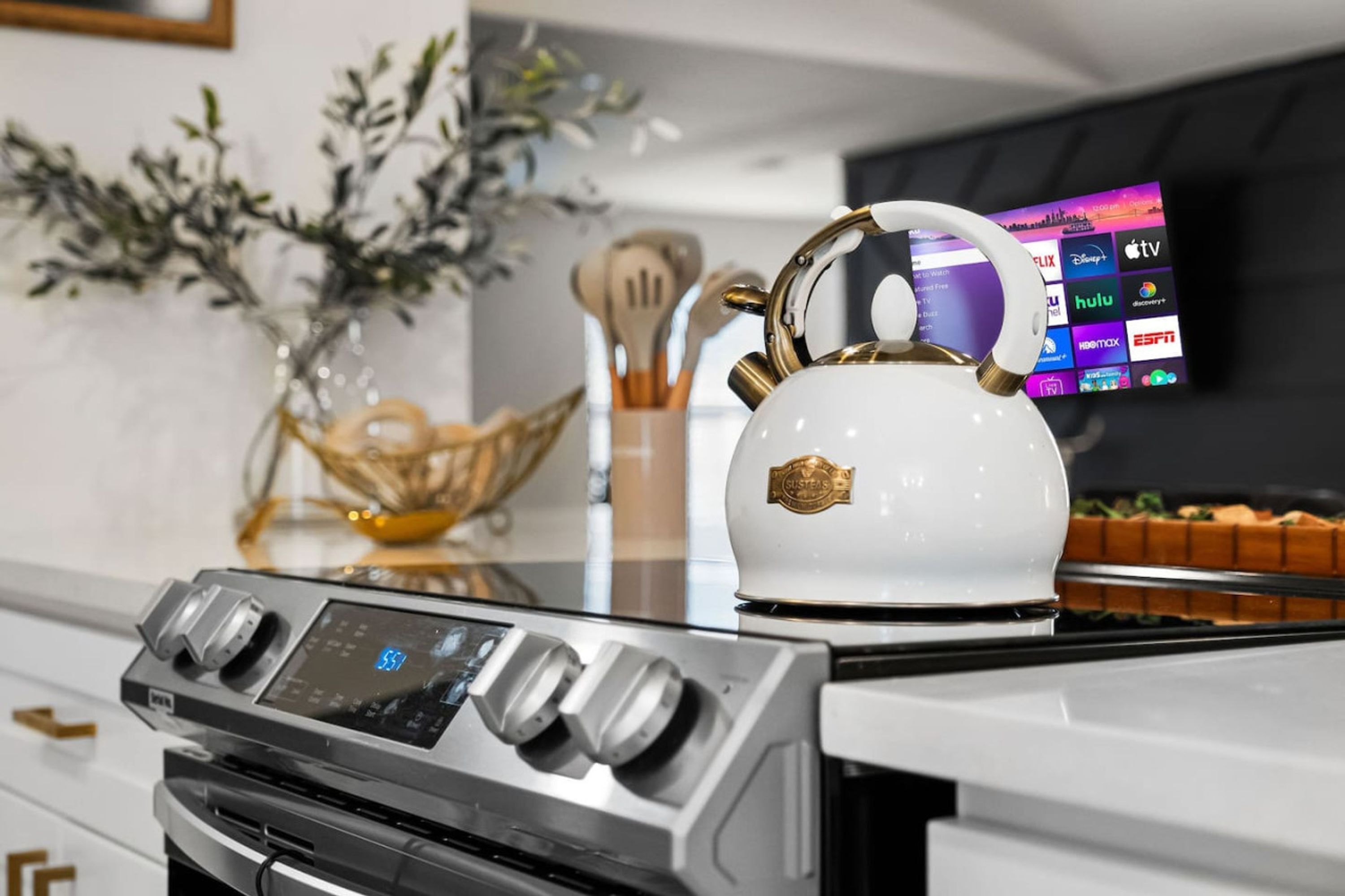 Une cuisine avec une bouilloire blanche sur la cuisinière, une horloge numérique et un téléviseur à écran plat affichant différentes chaînes.