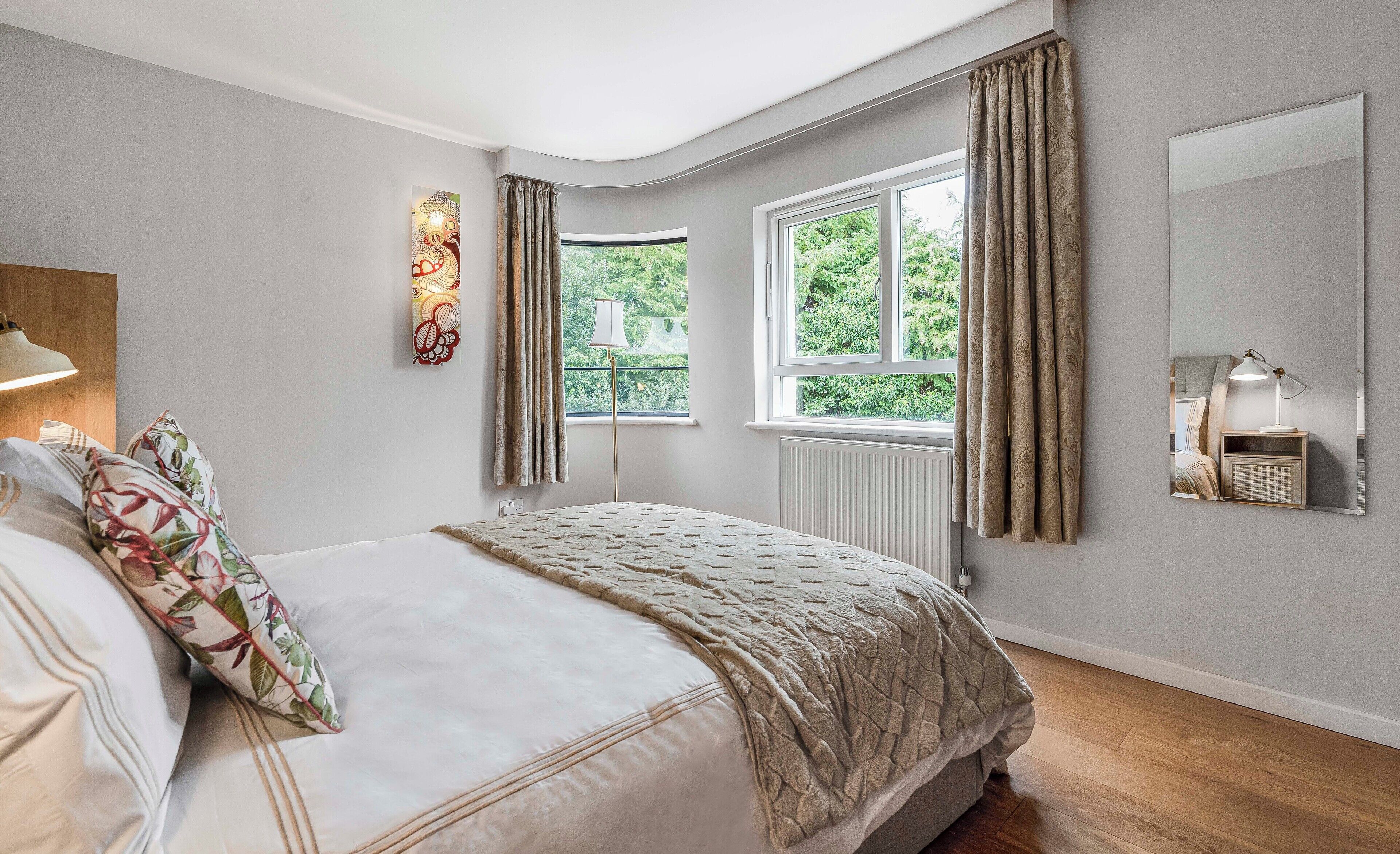 Une chambre à coucher comprenant un lit, une lampe de chevet, une fenêtre avec des rideaux, un radiateur et un miroir fixé au mur.