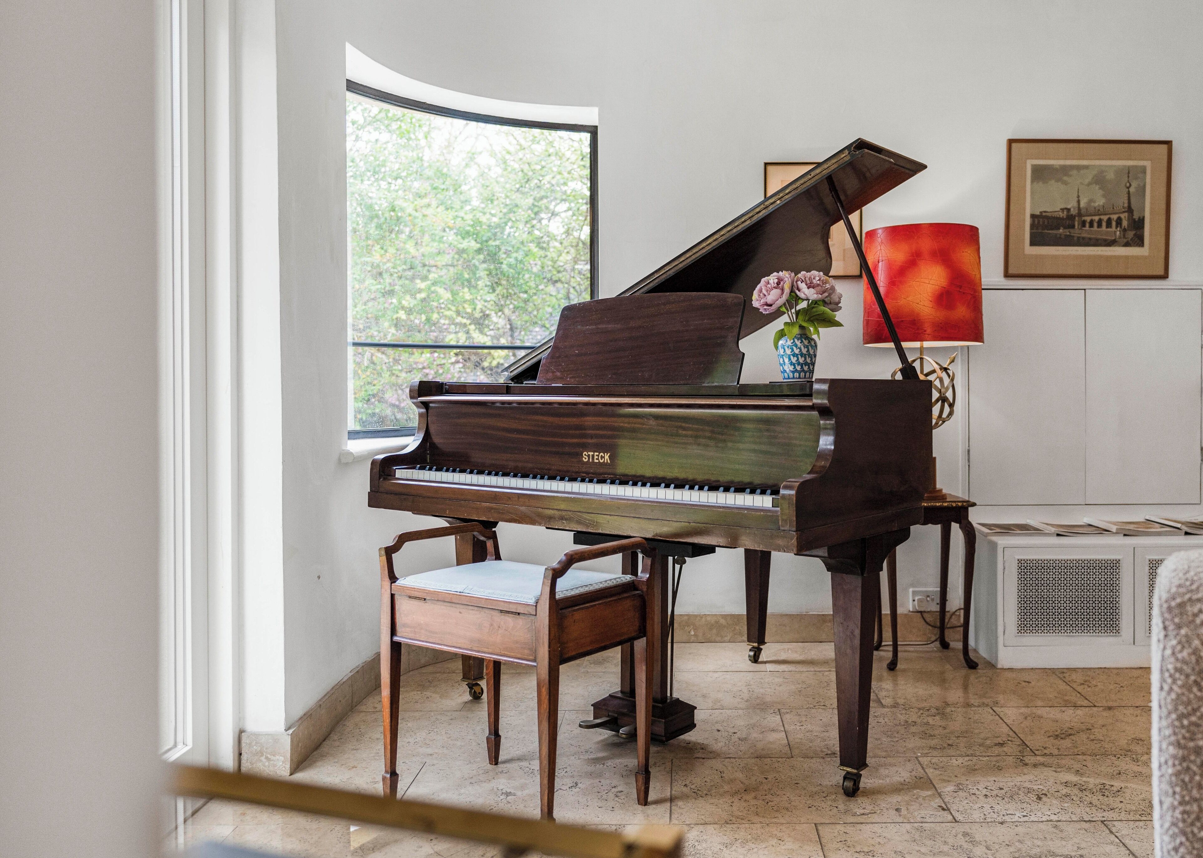 A grand piano in a room with a large window, a vase with flowers, and a framed picture on the wall.