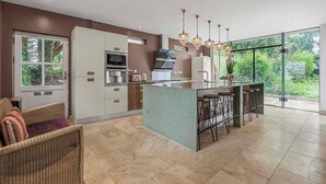 A modern kitchen with a central island, bar stools, and a view of greenery through large windows. - Art Deco Villa Oxford (Oxford)