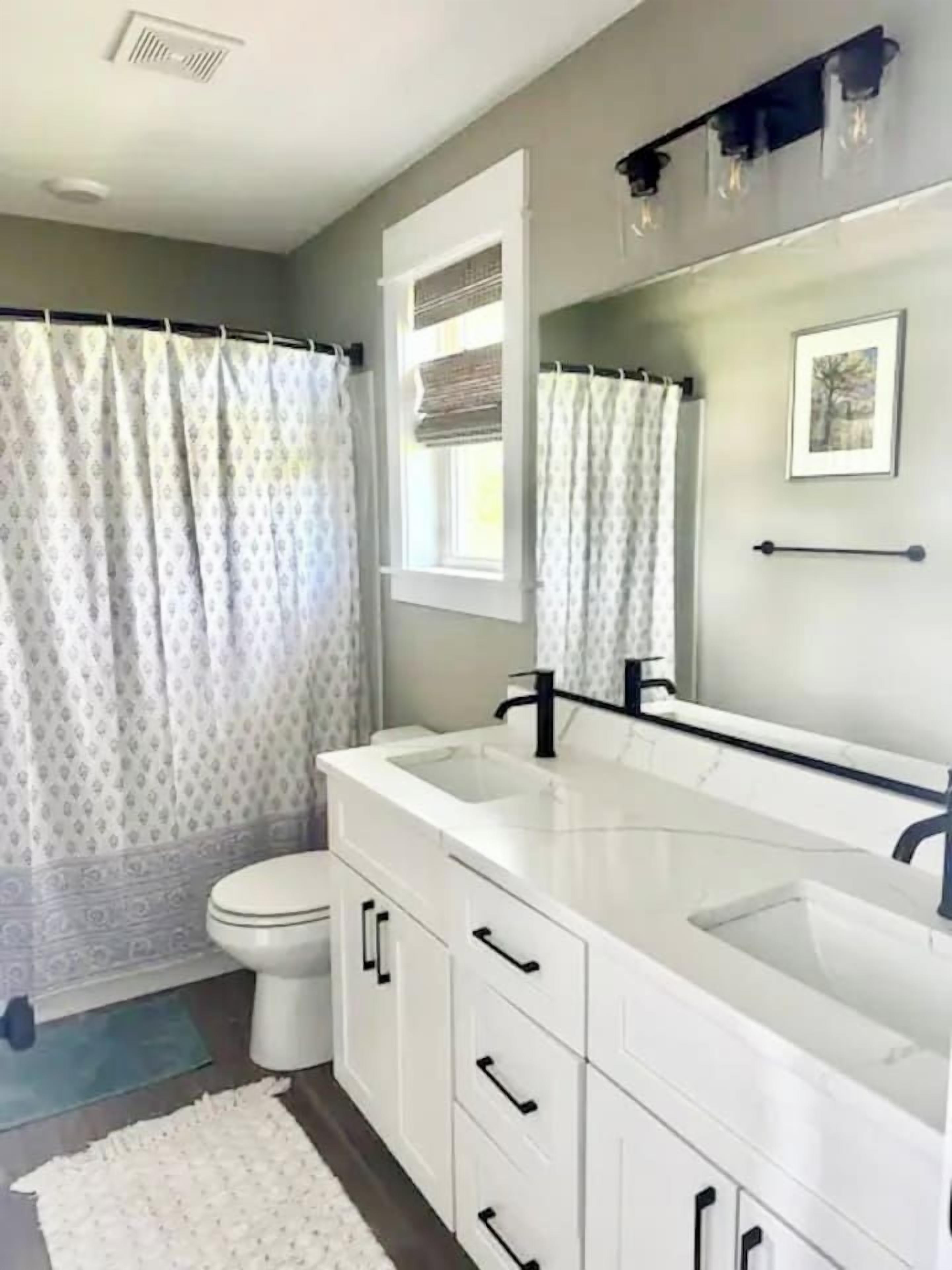 A bathroom with a white vanity, a toilet, a shower curtain, and a window with blinds.