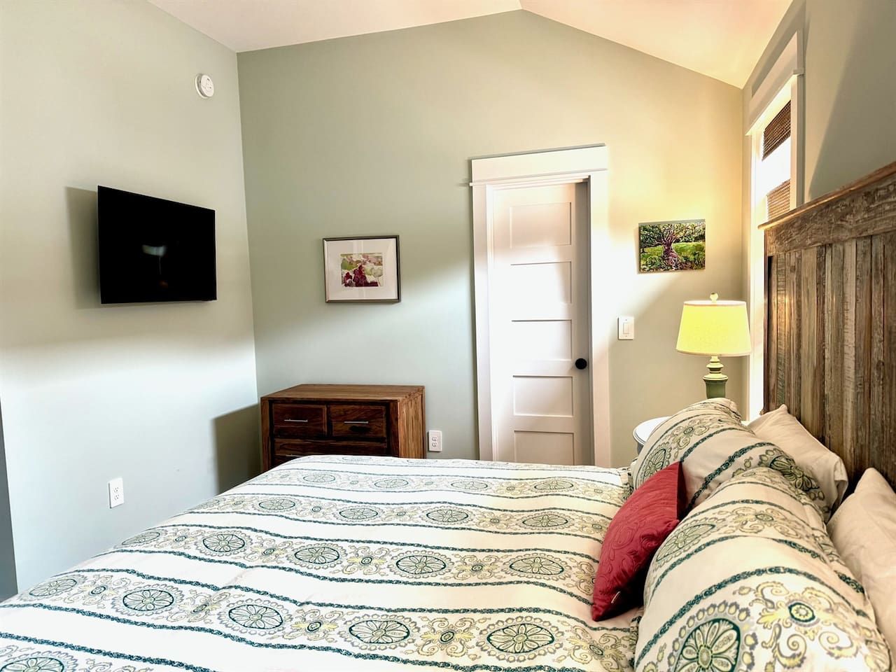 A bedroom with a bed, a wooden nightstand, a television mounted on the wall, and a door.