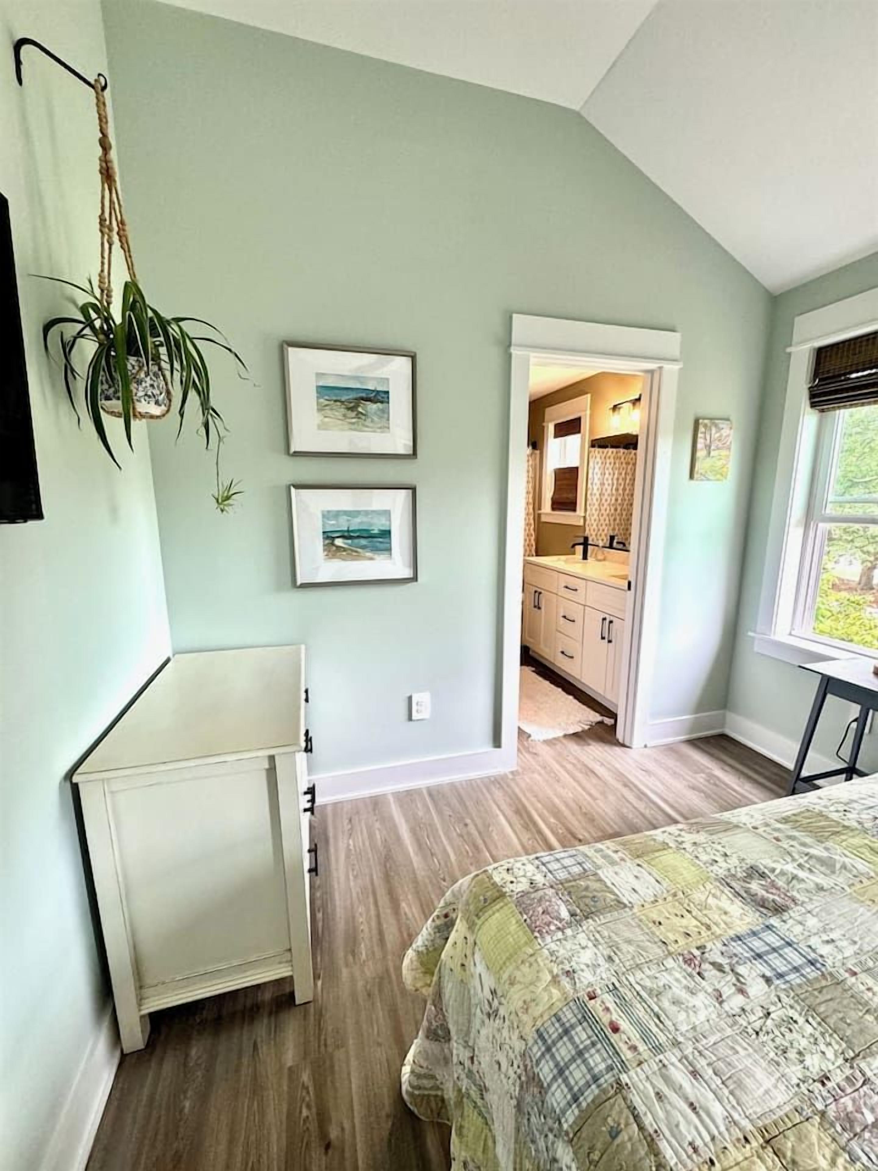 Une chambre à coucher avec un lit, une commode, une plante suspendue et une vue sur la salle de bain.