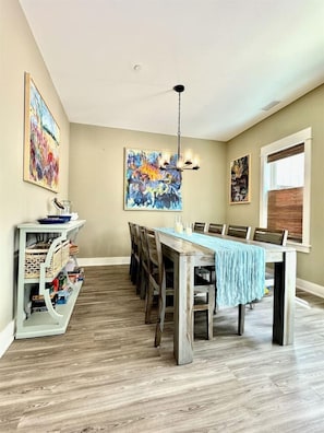 A dining area with a long table, chairs, a chandelier, and artwork on the walls. - NEW! Prime Location @Beach Club -Tranquil Seascape (Corolla)