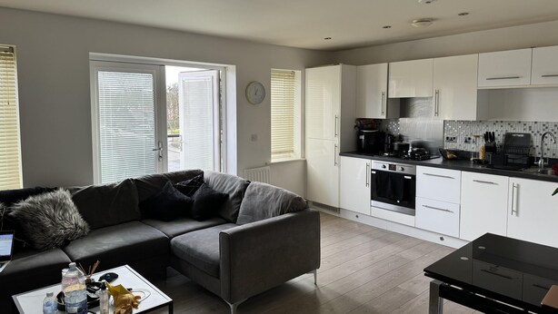 A modern living room with a grey sofa, a coffee table with various items, and a kitchen area with white cabinets and a black countertop. - Inviting 2-bed Apartment in Bushey (Bushey)